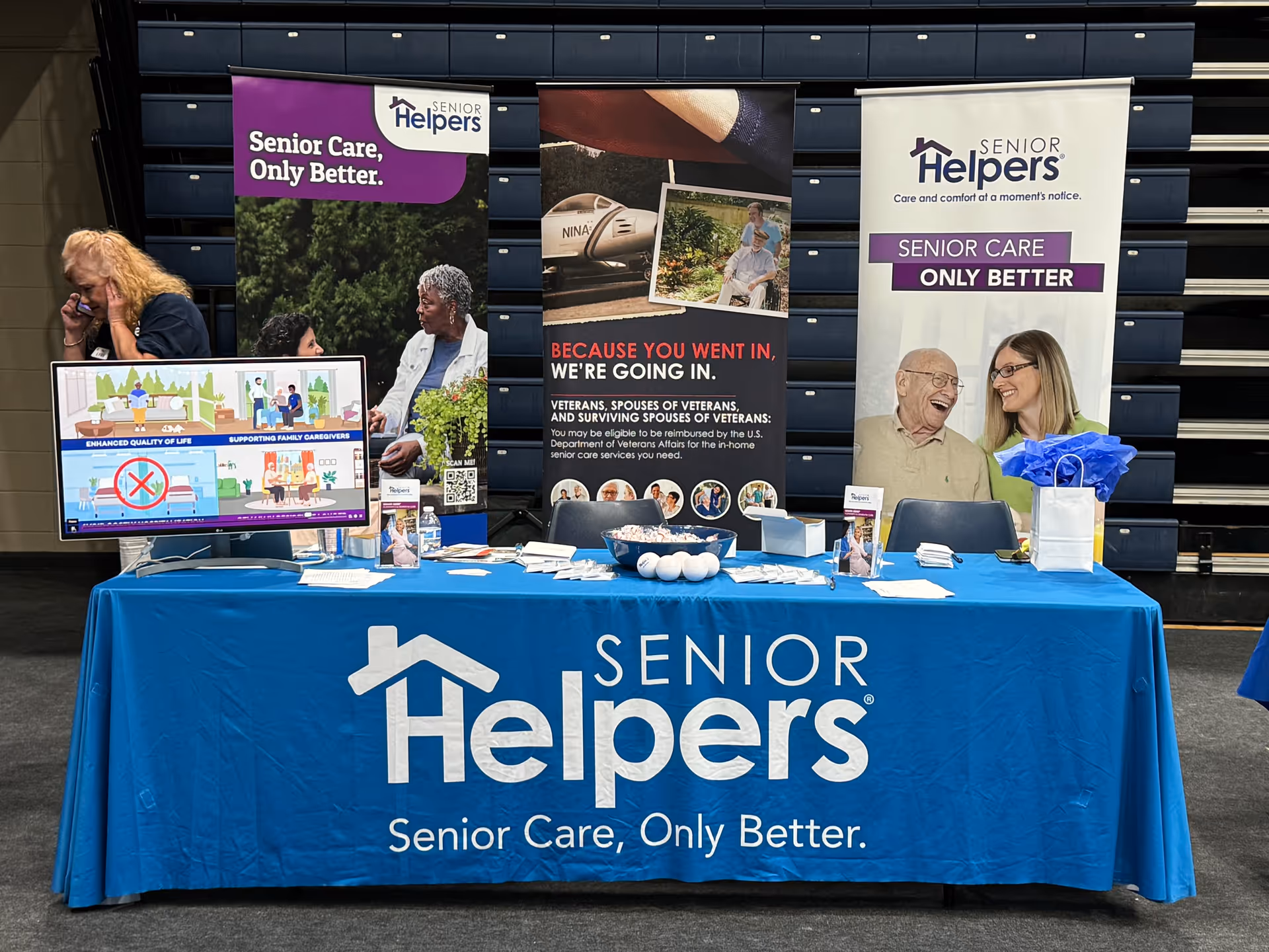 Promotional display table with Senior Helpers banners in an indoor hall.