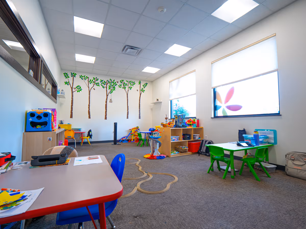 Bright children's playroom with colorful tables, chairs, toys, and a tree mural on the wall.