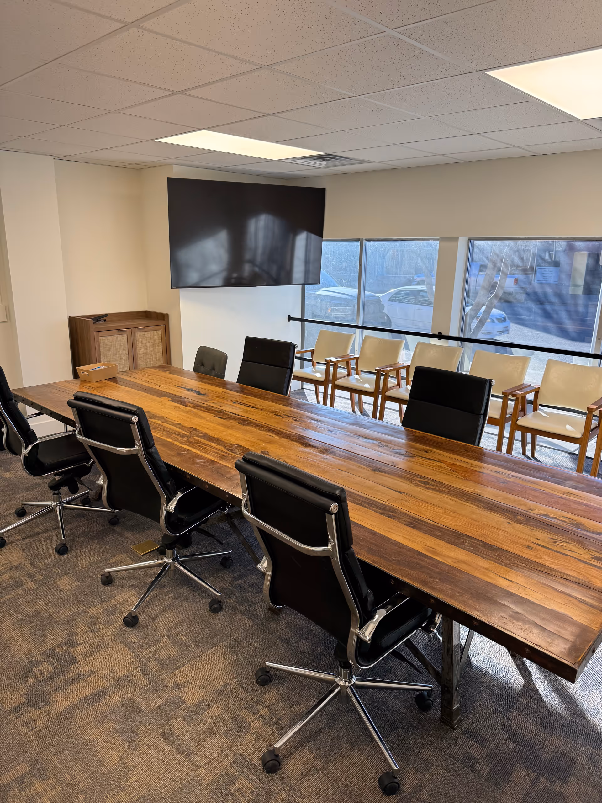A conference room with a long wooden table, black chairs, and a wall-mounted screen.