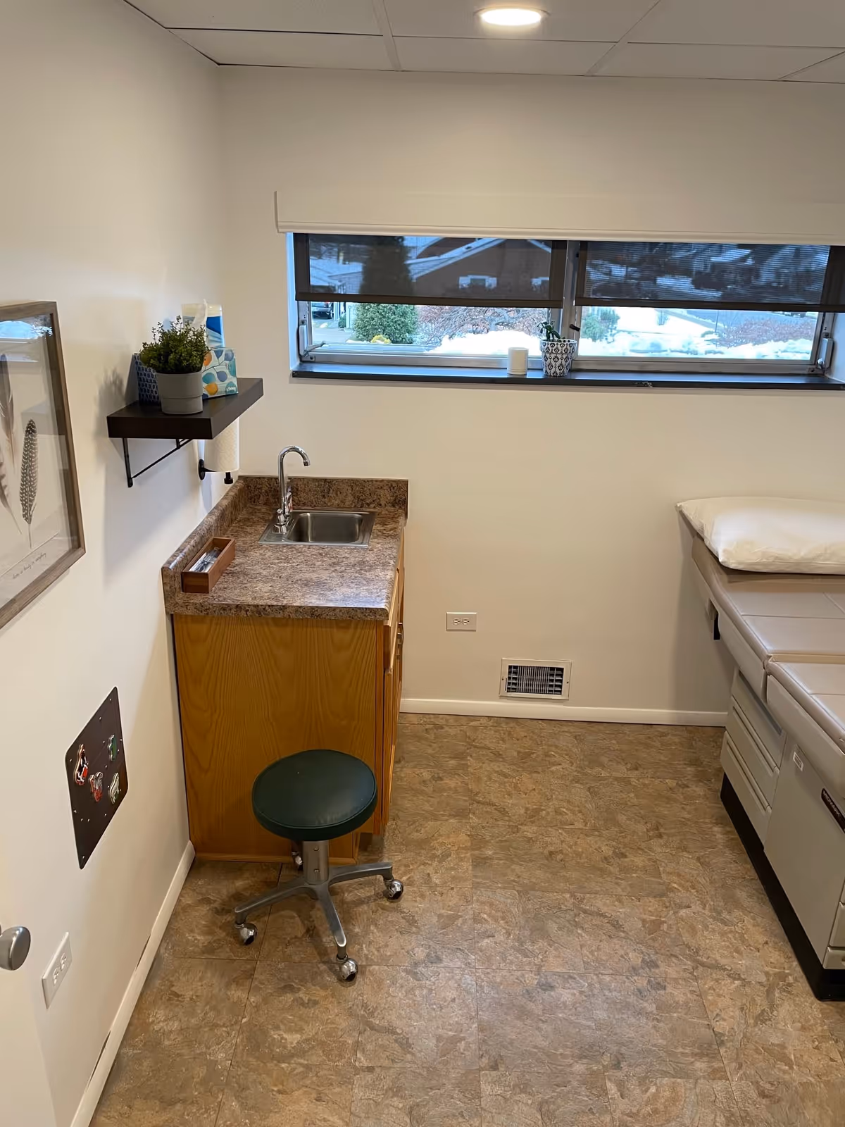 Small medical examination room with sink, counter, stool, window, and examination bed.