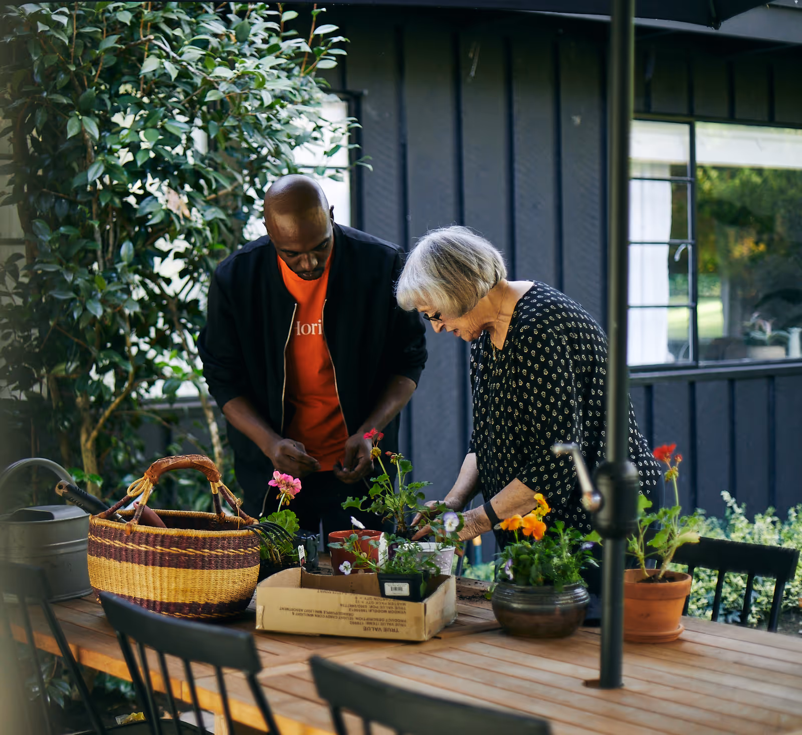 Two people pot flowers at an outdoor wooden table on a sunny patio