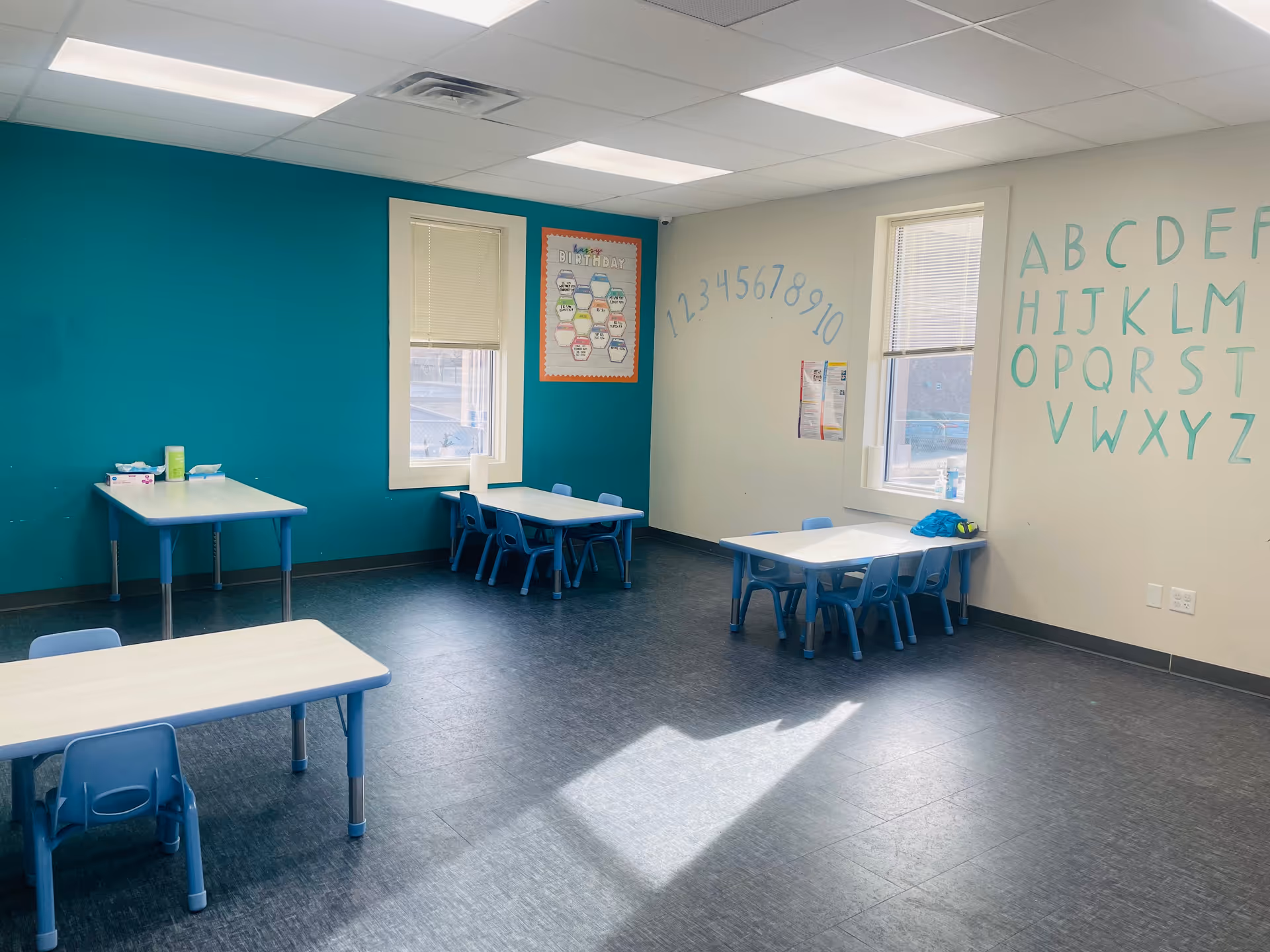 Bright classroom-style therapy room with small blue tables, chairs, and alphabet mural.