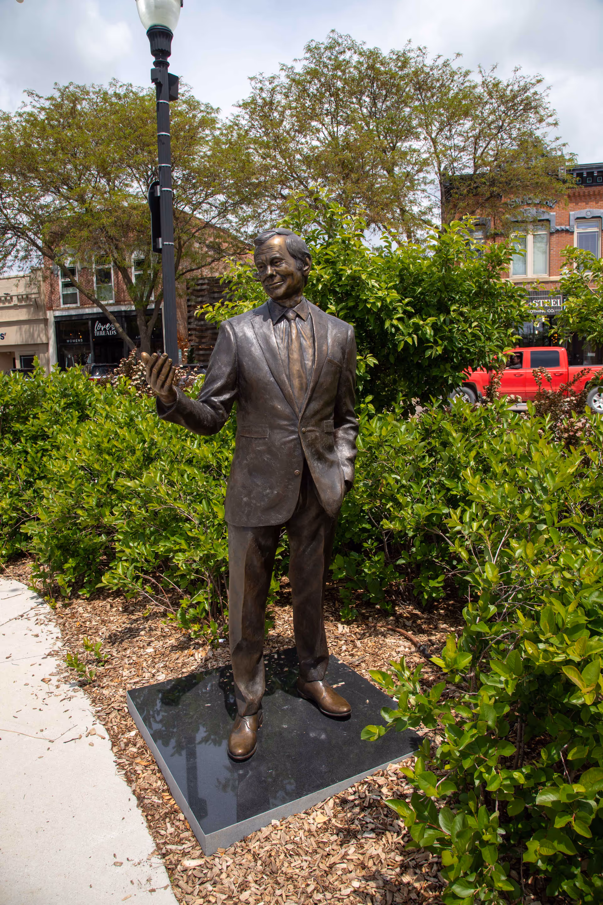 Bronze statue of a man in a suit stands on a pedestal amid greenery outdoors.