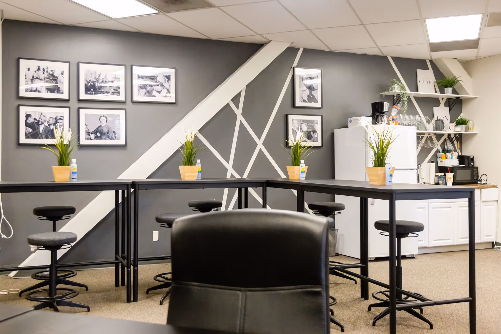 Modern staff kitchenette with high tables, stools, coffee station, plants, and framed photos.
