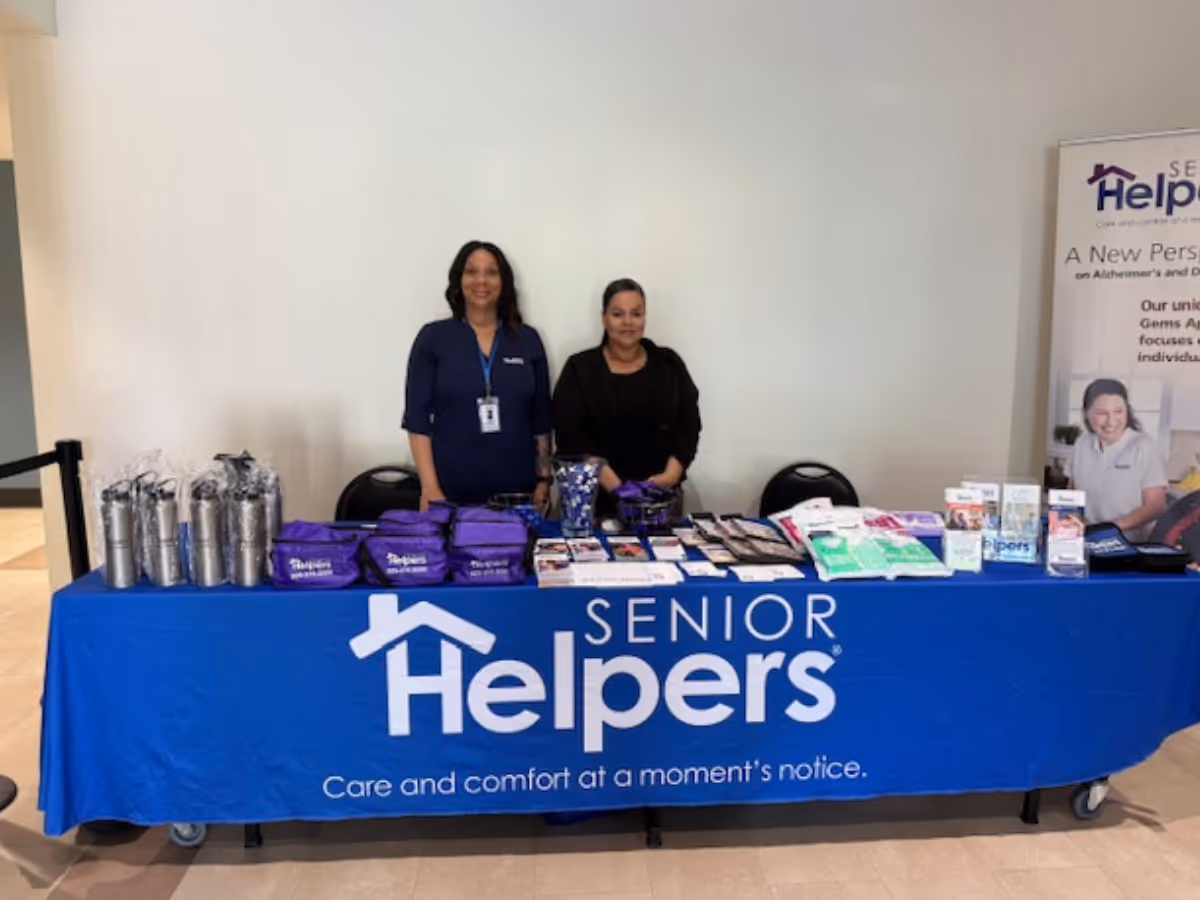 Two staff members stand behind a blue Senior Helpers information table with brochures.