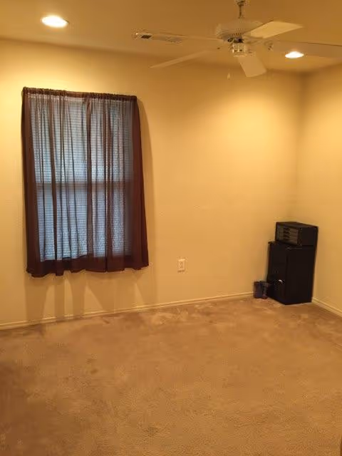 Empty beige living room with window, brown curtain, ceiling fan, and black stereo cabinet.
