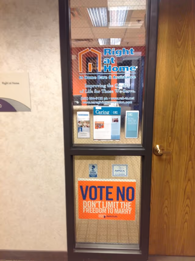 In a hallway, a glass door displays Right at Home signage and a Vote No poster.