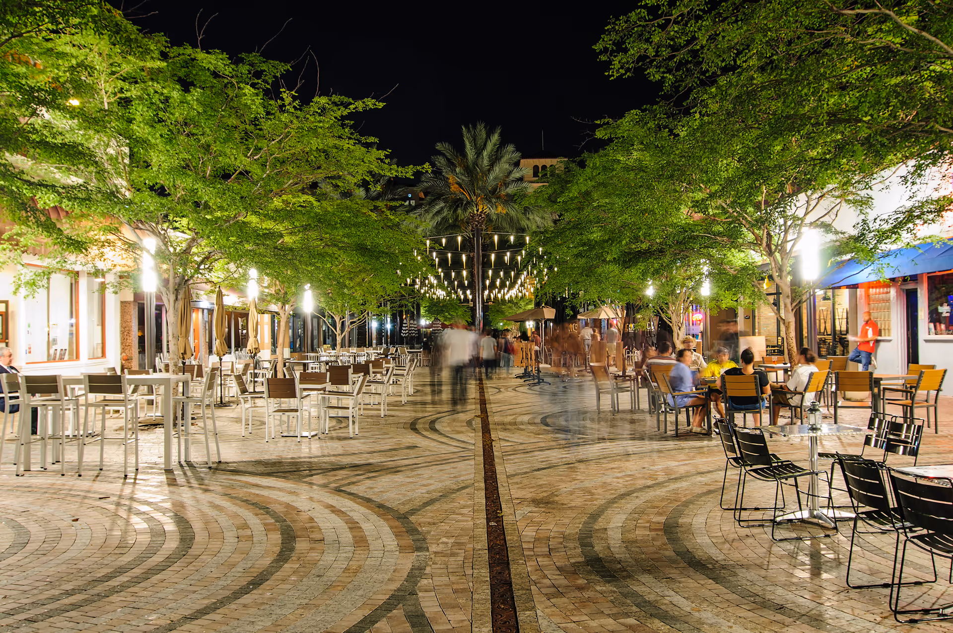 Night view of an outdoor plaza with string lights, trees, and many tables and chairs.