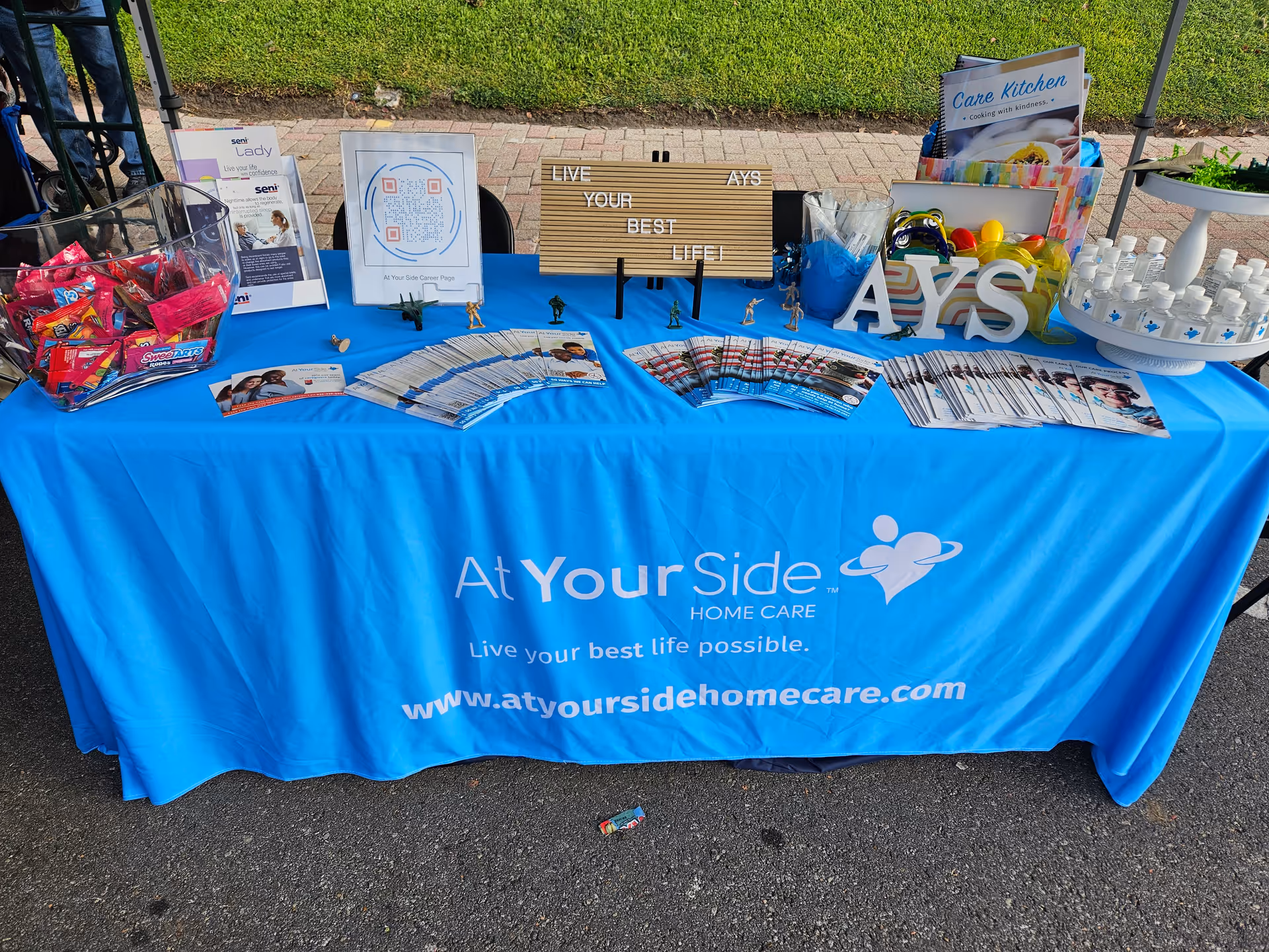 Outdoor promotional booth with blue At Your Side Home Care tablecloth, brochures, and signage.
