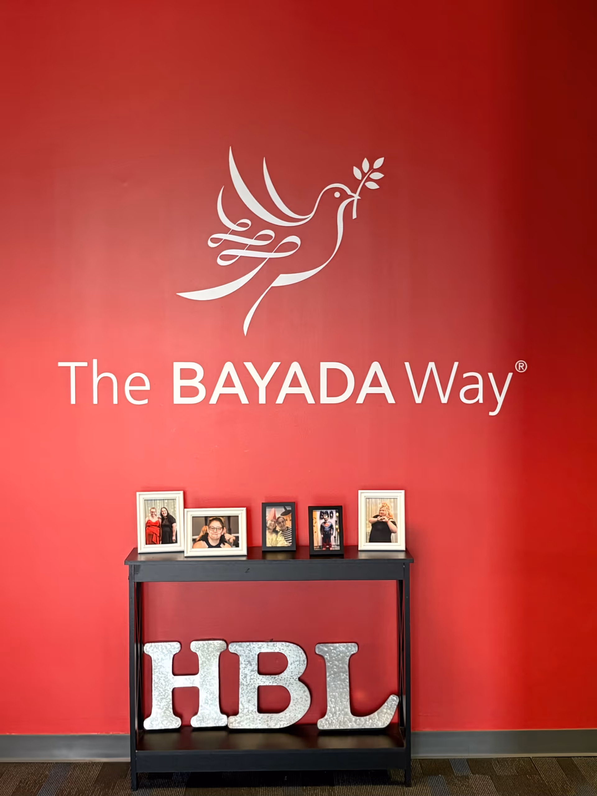Red wall with The BAYADA Way logo and framed photos on a black console table.