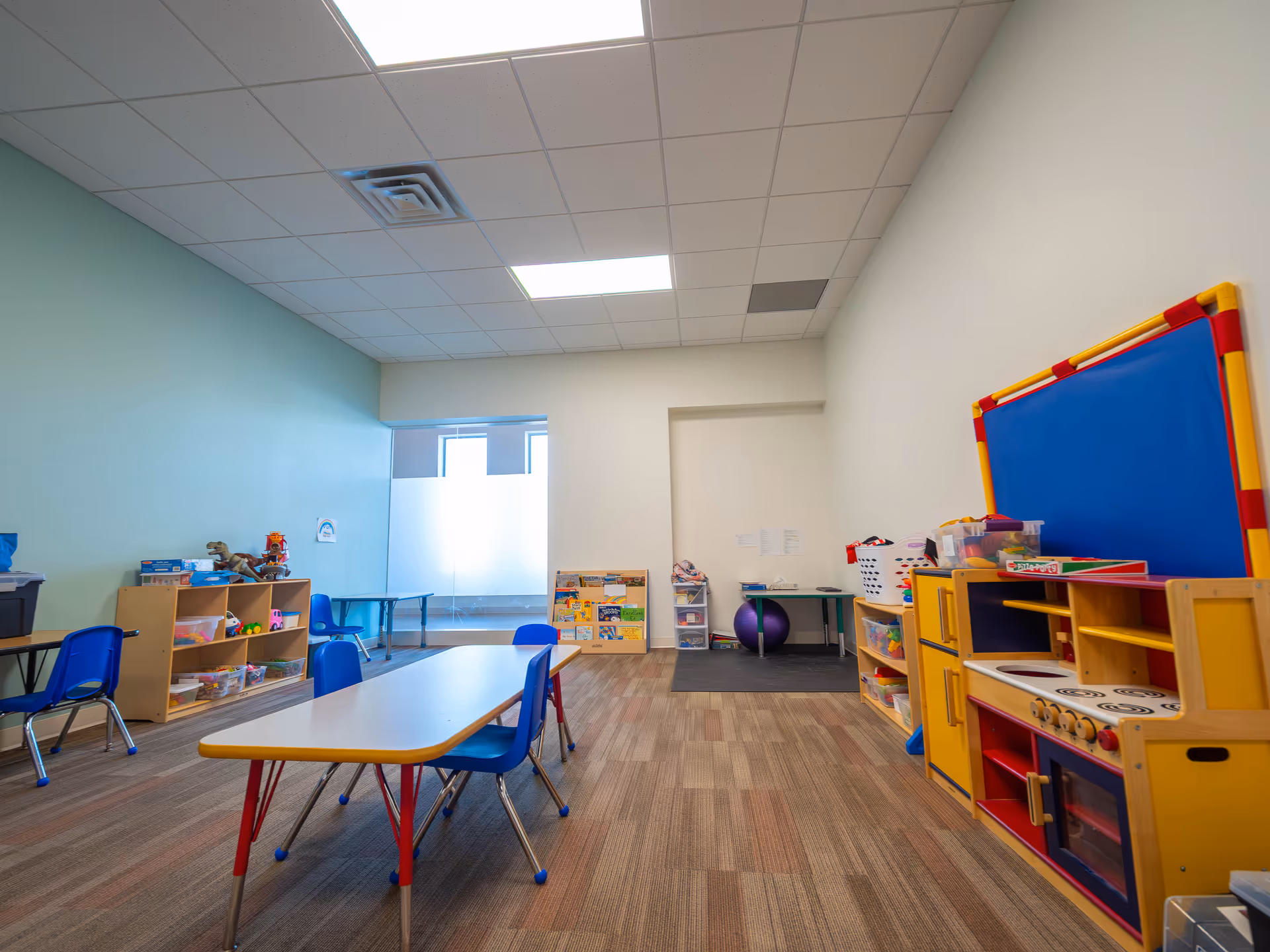 Bright children's activity room with small tables, blue chairs, and a colorful play kitchen.