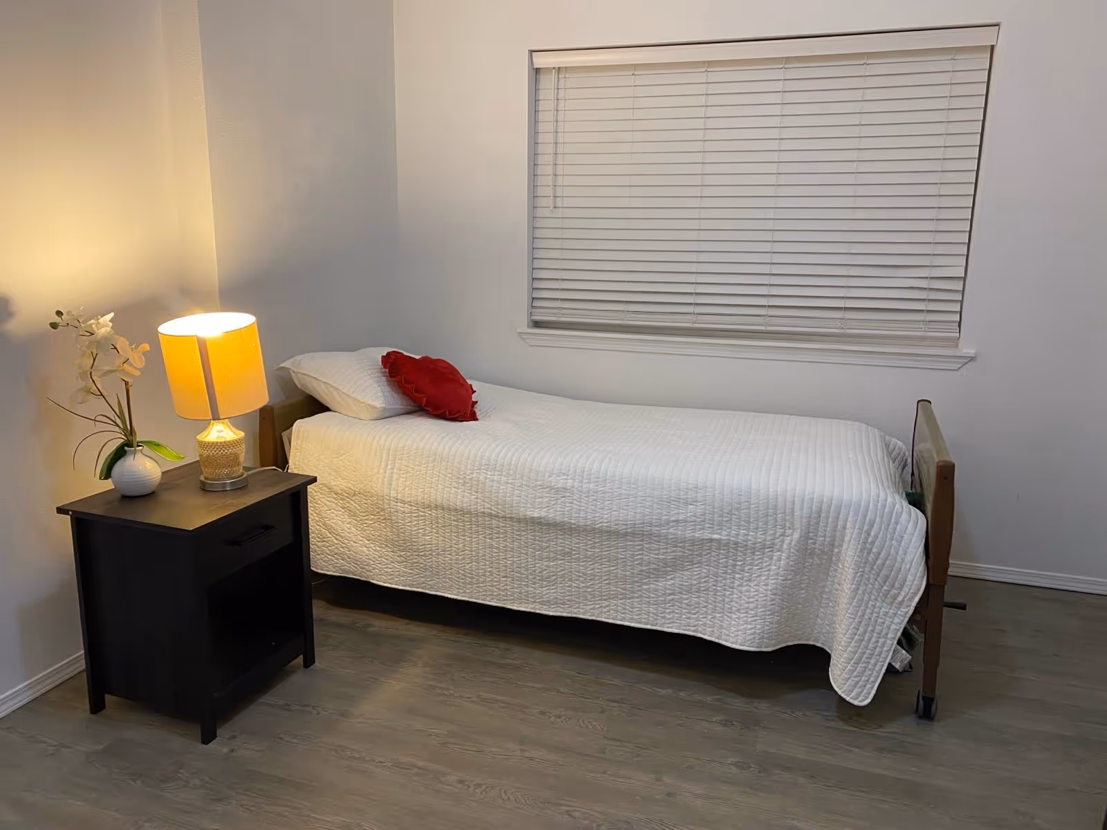 A small bedroom with a single bed, nightstand, lamp, and blinds.