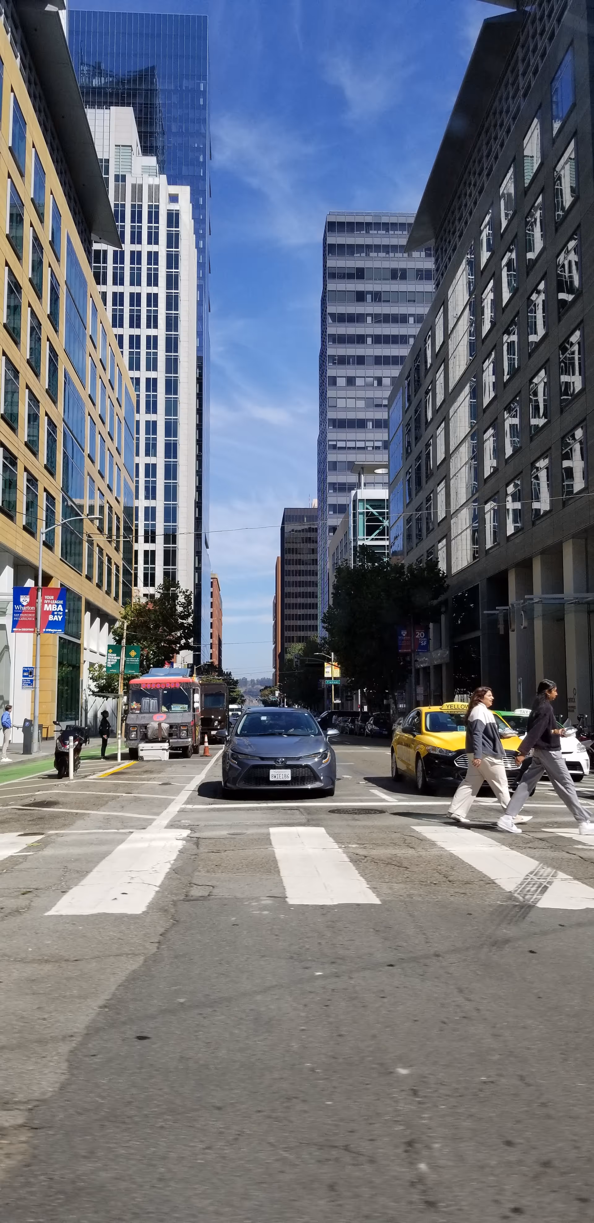 City street with tall office buildings, cars, and pedestrians at a crosswalk.