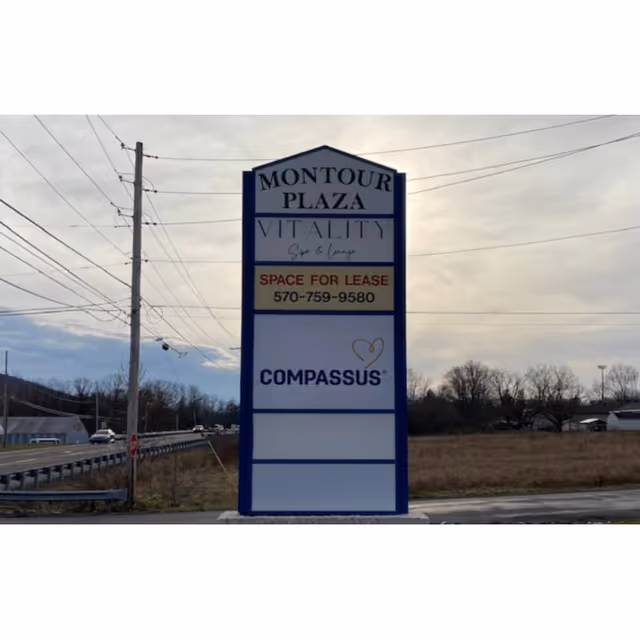 A tall blue-and-white roadside sign for Montour Plaza Vitality and Compassus near a road with lines.