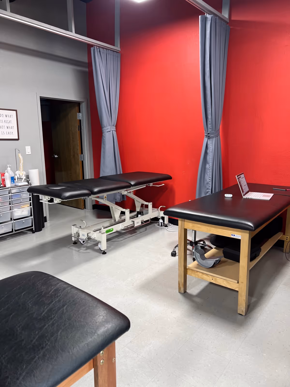 Therapy room with adjustable treatment table, red walls, blue curtains, and a laptop on a desk