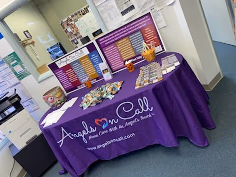 Purple tablecloth information table with brochures and Angels On Call signage in a hallway.