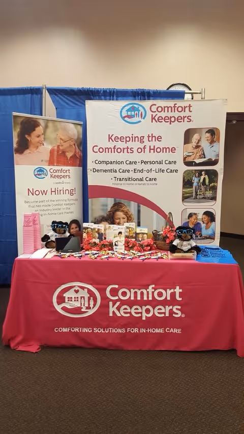 Indoor exhibition booth for Comfort Keepers with banners, pink tablecloth, and staff.