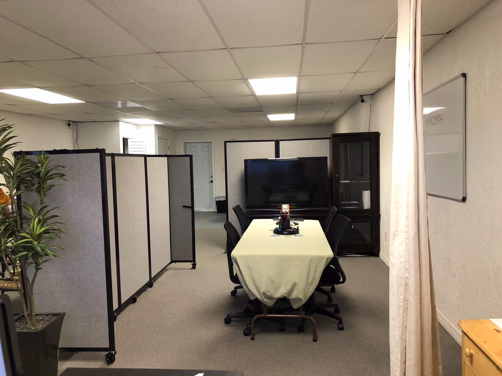 Meeting room with table, chairs, large TV, privacy screens, plant, and whiteboard.