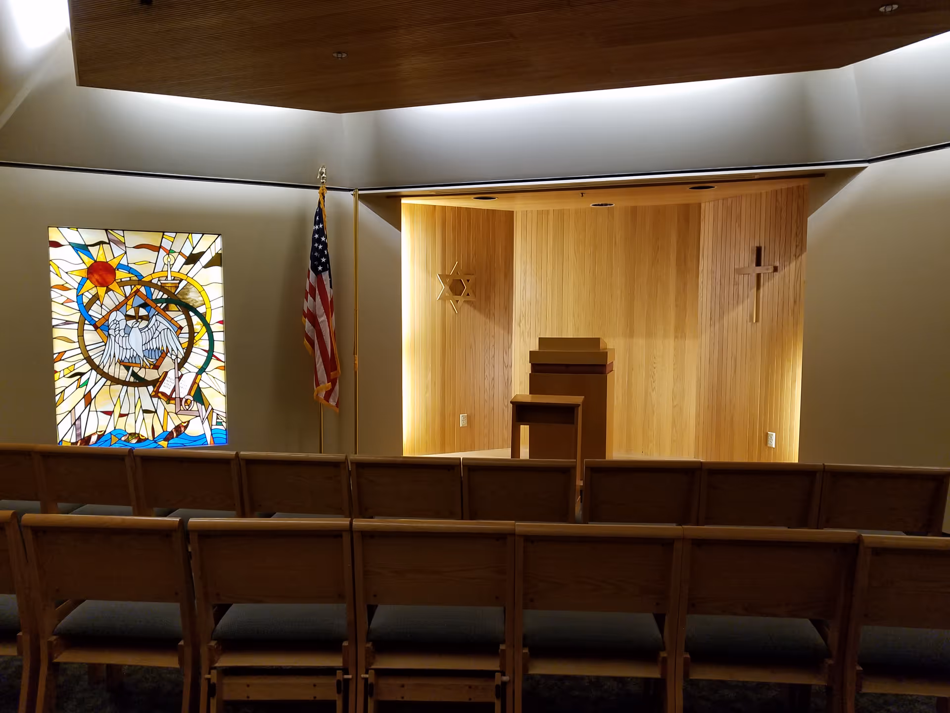 Chapel interior with wooden altar backdrop, lectern, cross, stained glass window, flag, and pews.