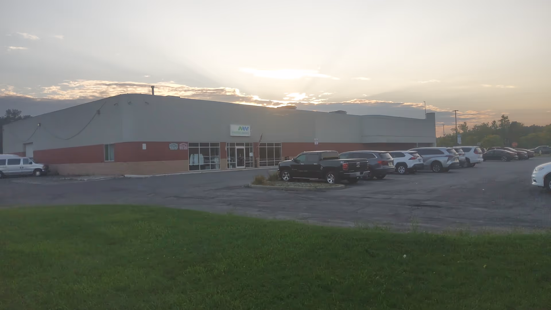 A single-story building with a parking lot in front, at sunset, under a cloudy sky.