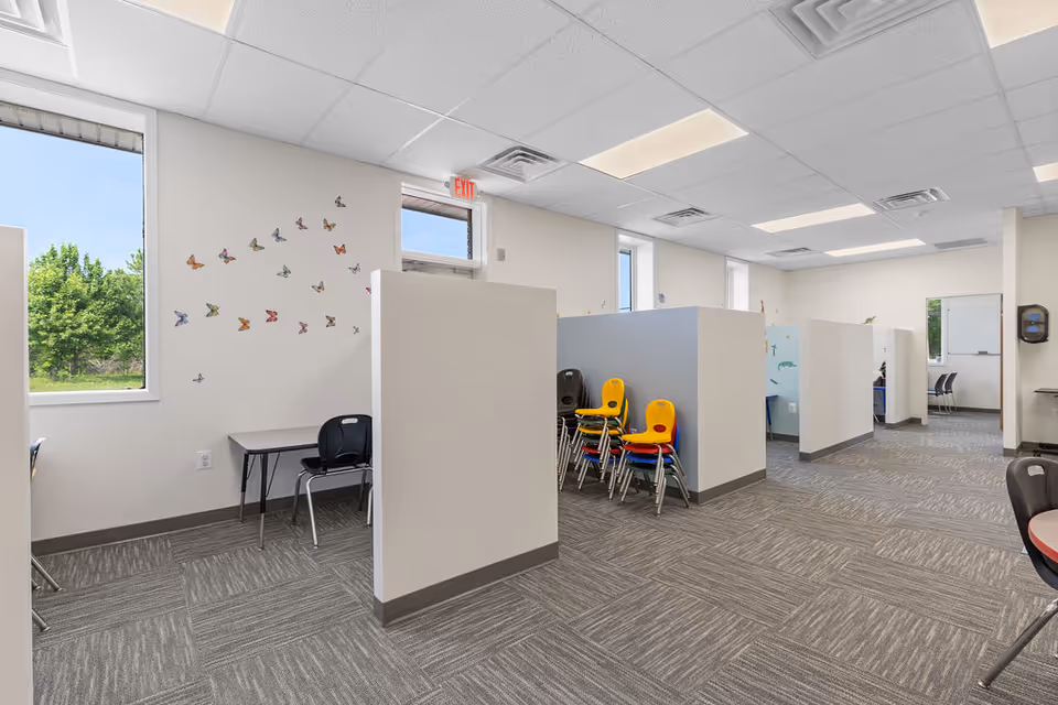Interior therapy space with partitioned cubicles, stacked chairs, butterfly decals, and large windows.