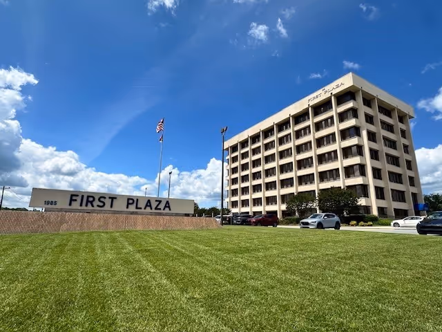Exterior view of a multi-story building with a large 'FIRST PLAZA' sign and parked cars