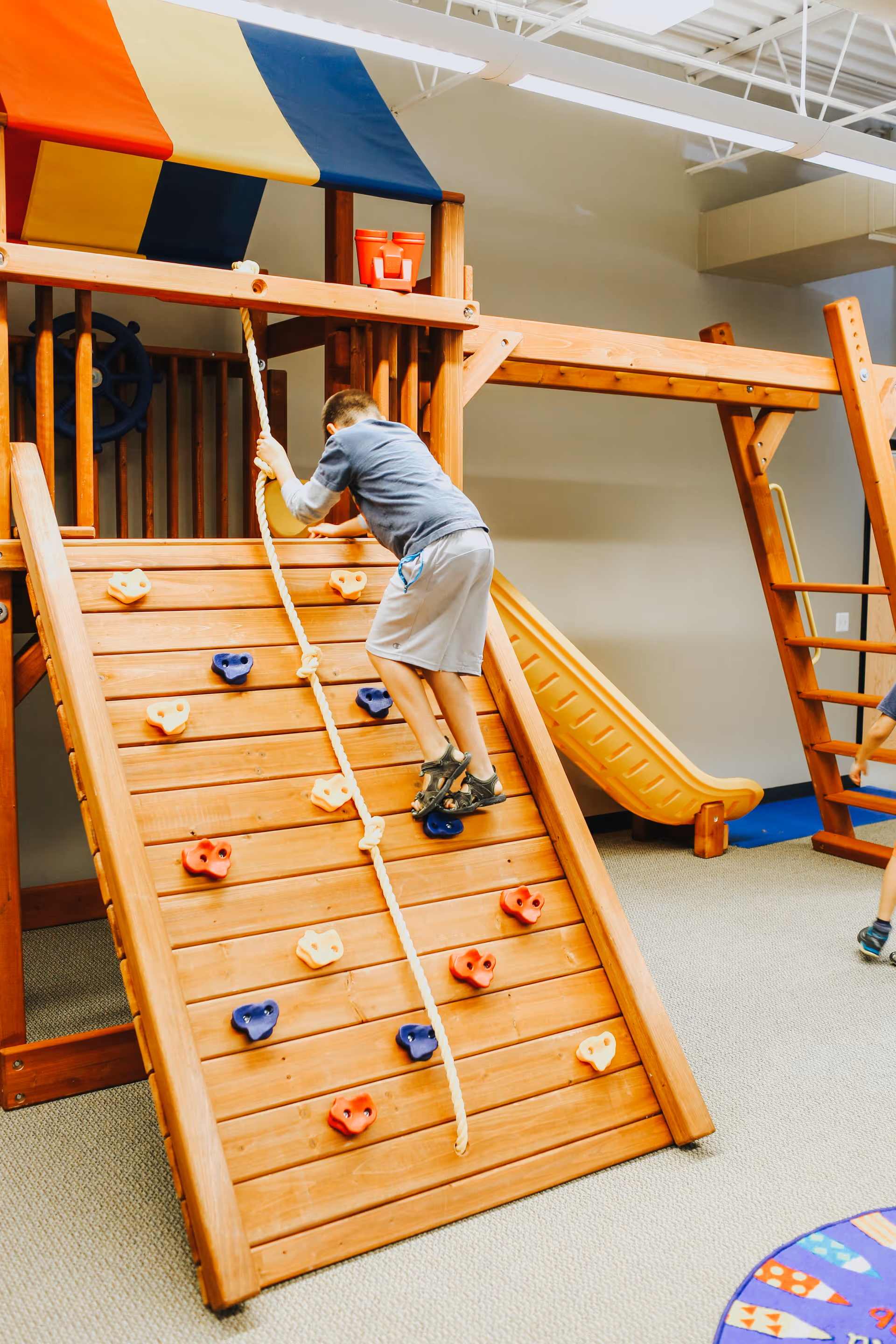 A person climbs a wooden indoor climbing wall in a colorful play area.