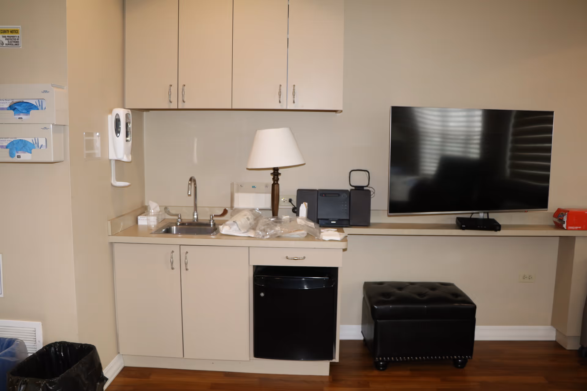 A compact kitchenette with sink, cabinets, mini fridge, lamp, and TV.