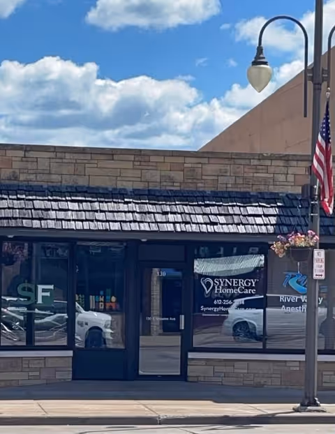 Storefront with Synergy HomeCare signage, blue door, stone facade, American flag on sidewalk.
