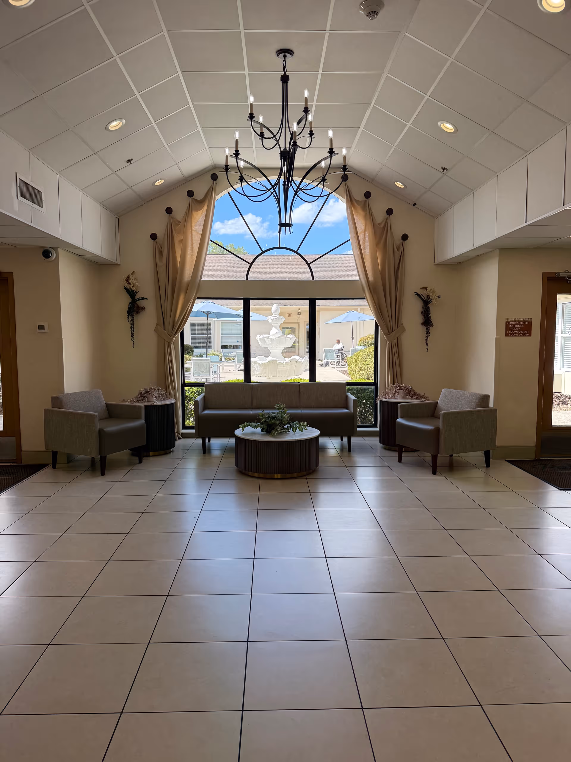 Bright lobby with beige seating, central coffee table, chandelier, and large arched window.