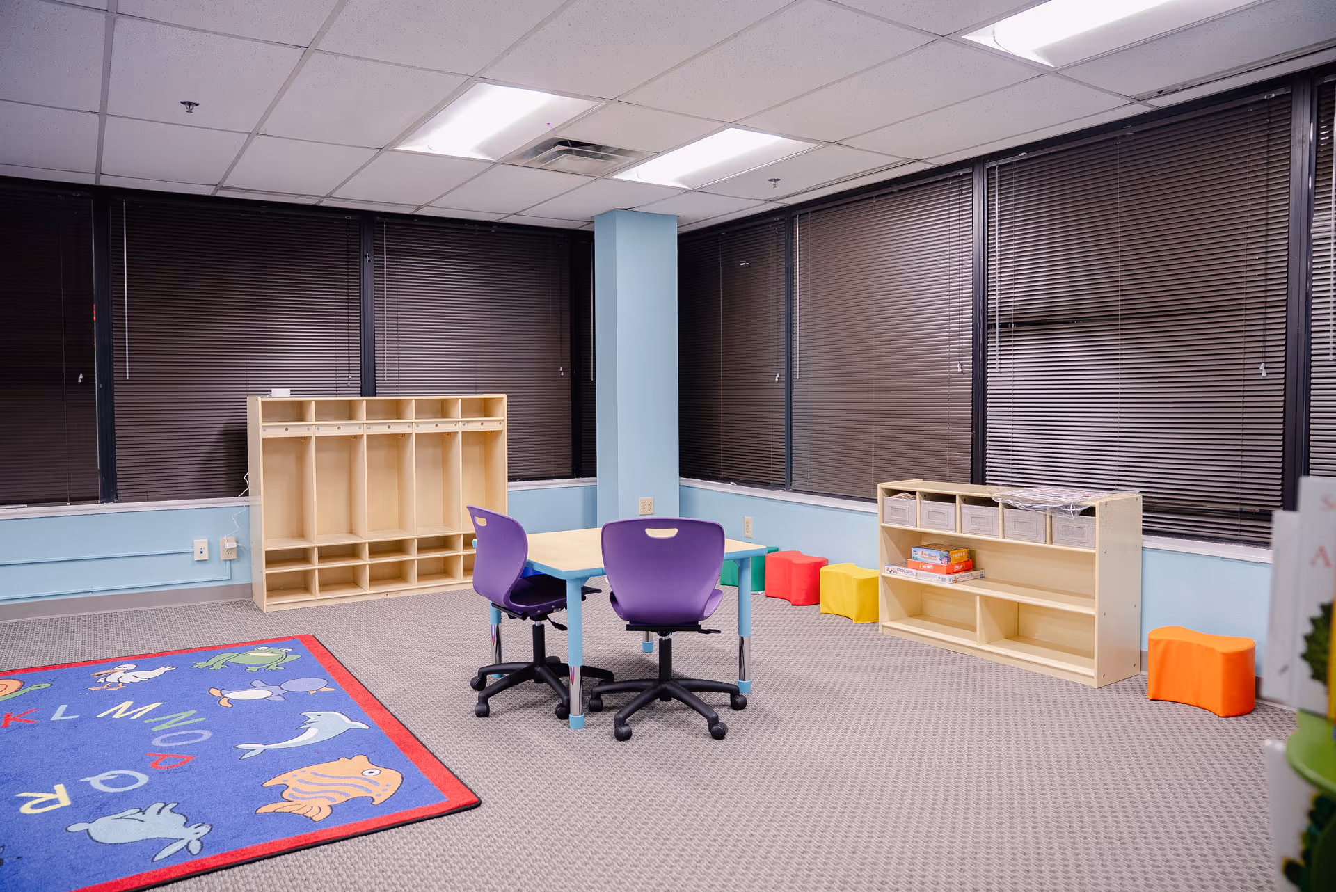 Bright activity room with small table, purple chairs, shelves, and a colorful rug.