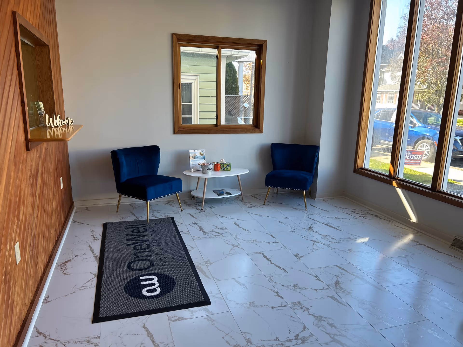 Bright lobby seating area with two blue chairs, a round table, and large windows.