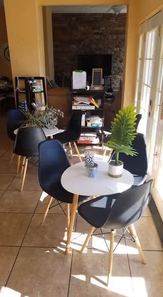 Light-filled lounge area with round white tables, black chairs, plants, and a TV.