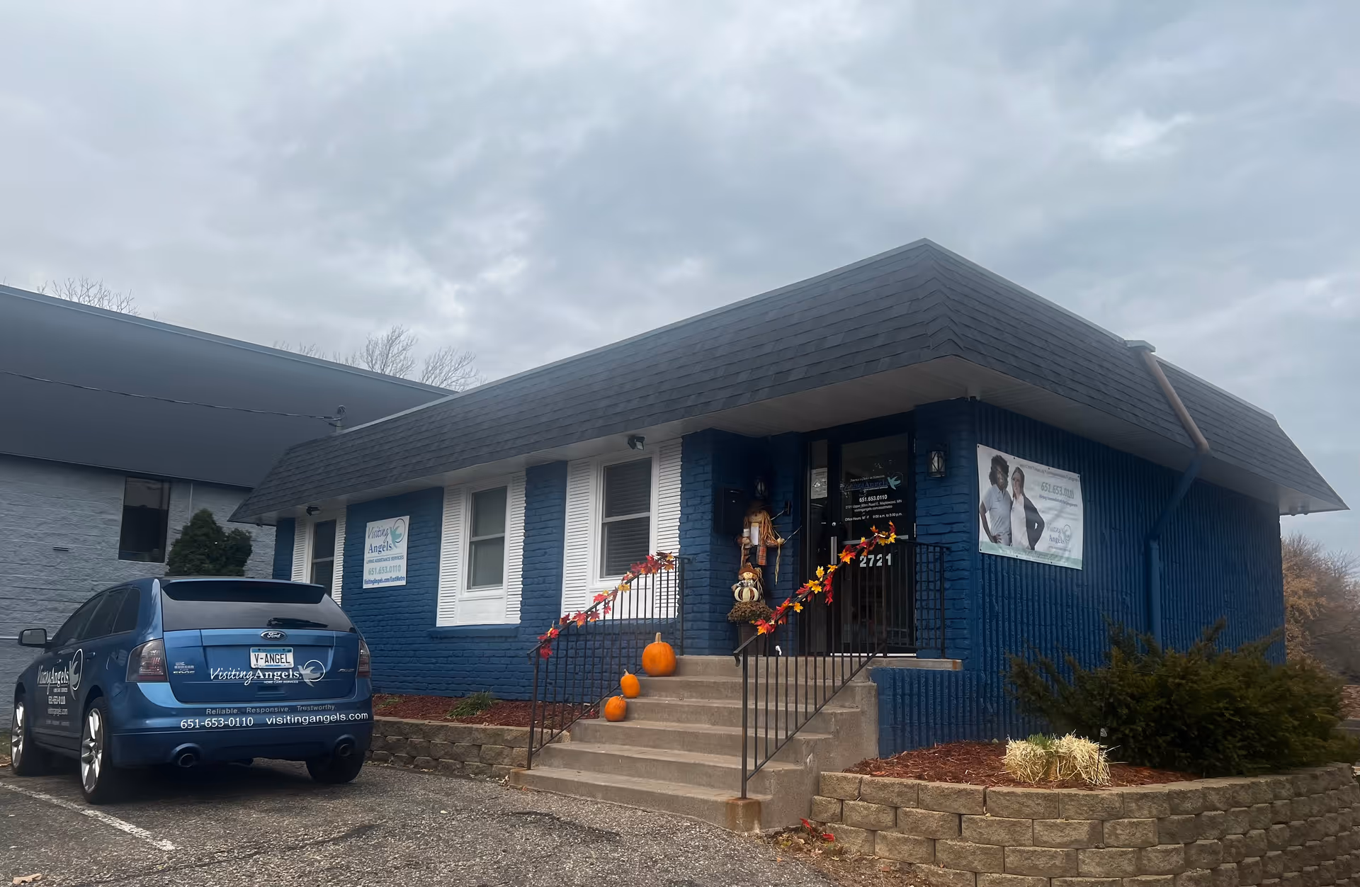 Blue brick Visiting Angels storefront exterior with pumpkins on steps and a parked branded car.