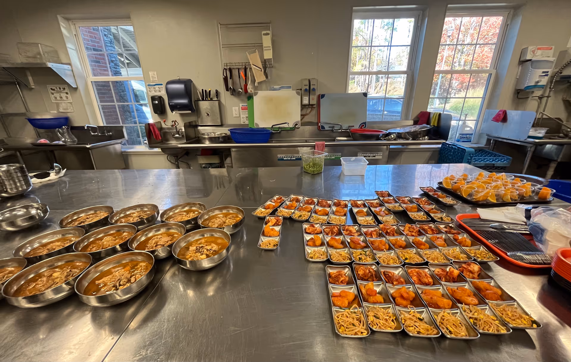 Commercial-style kitchen with stainless steel counter and many trays of prepared meals.