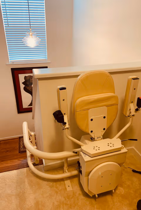 Beige stairlift chair with rails mounted along wall inside a residential-style room.
