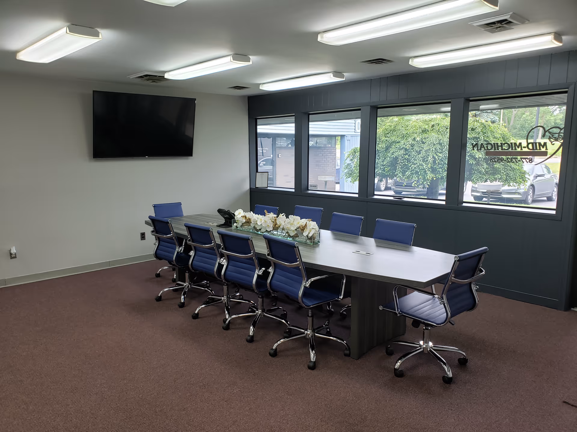 A conference room with a long table, blue chairs, and a wall-mounted TV.