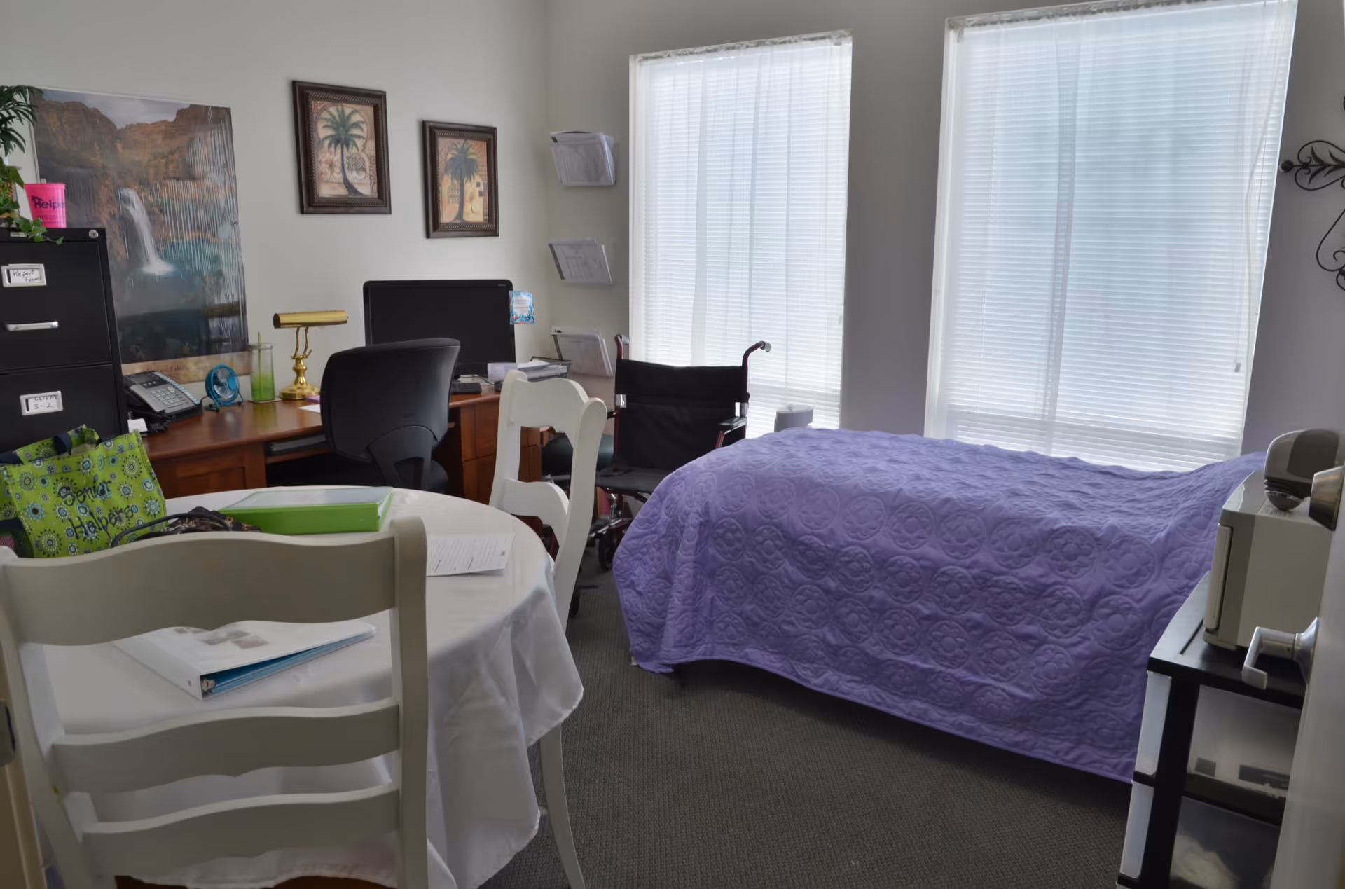 A furnished senior living room with a bed, round table, desk, and wheelchair.