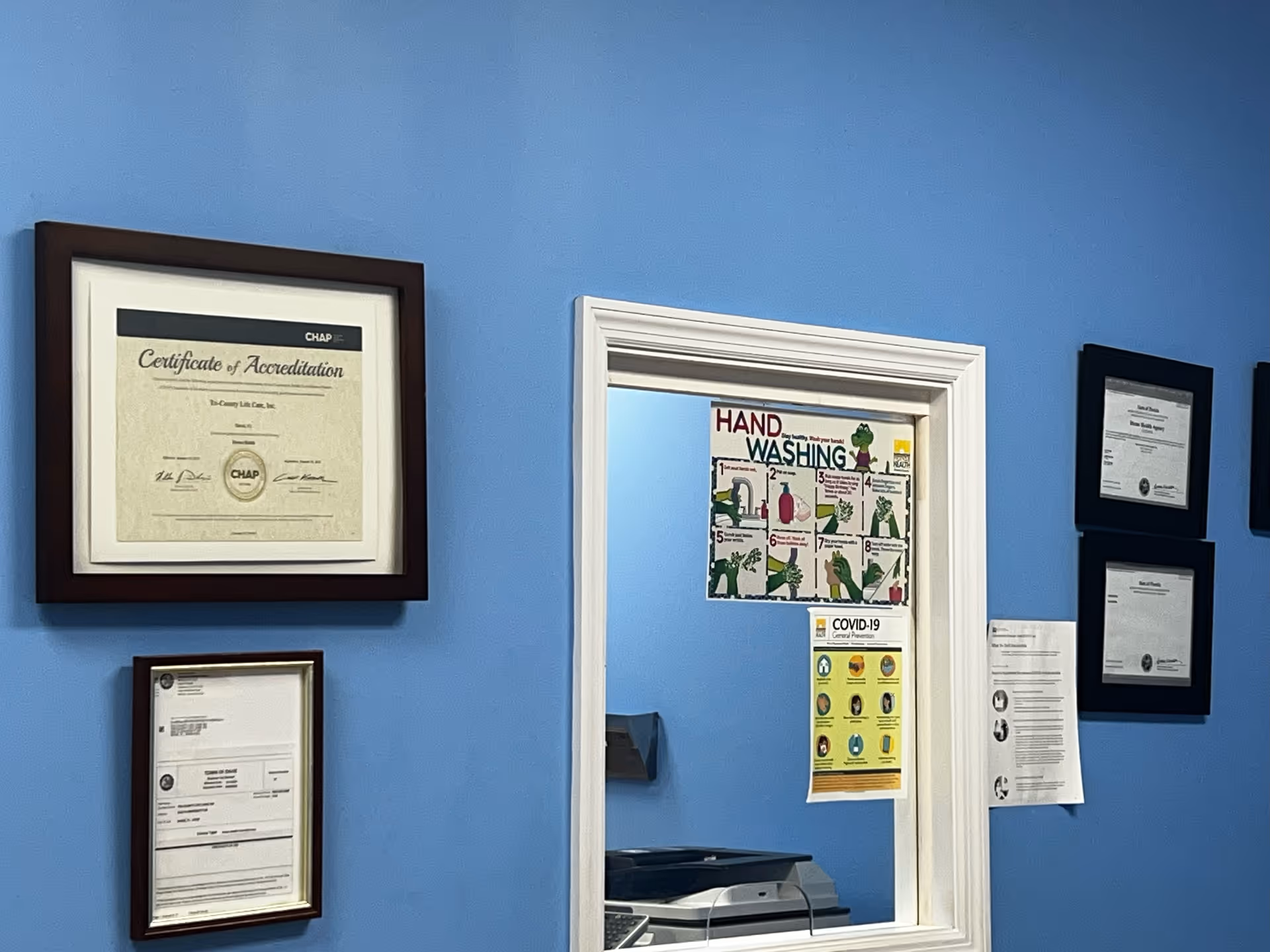 Blue hallway wall with framed certificates and a doorway displaying hand washing posters.