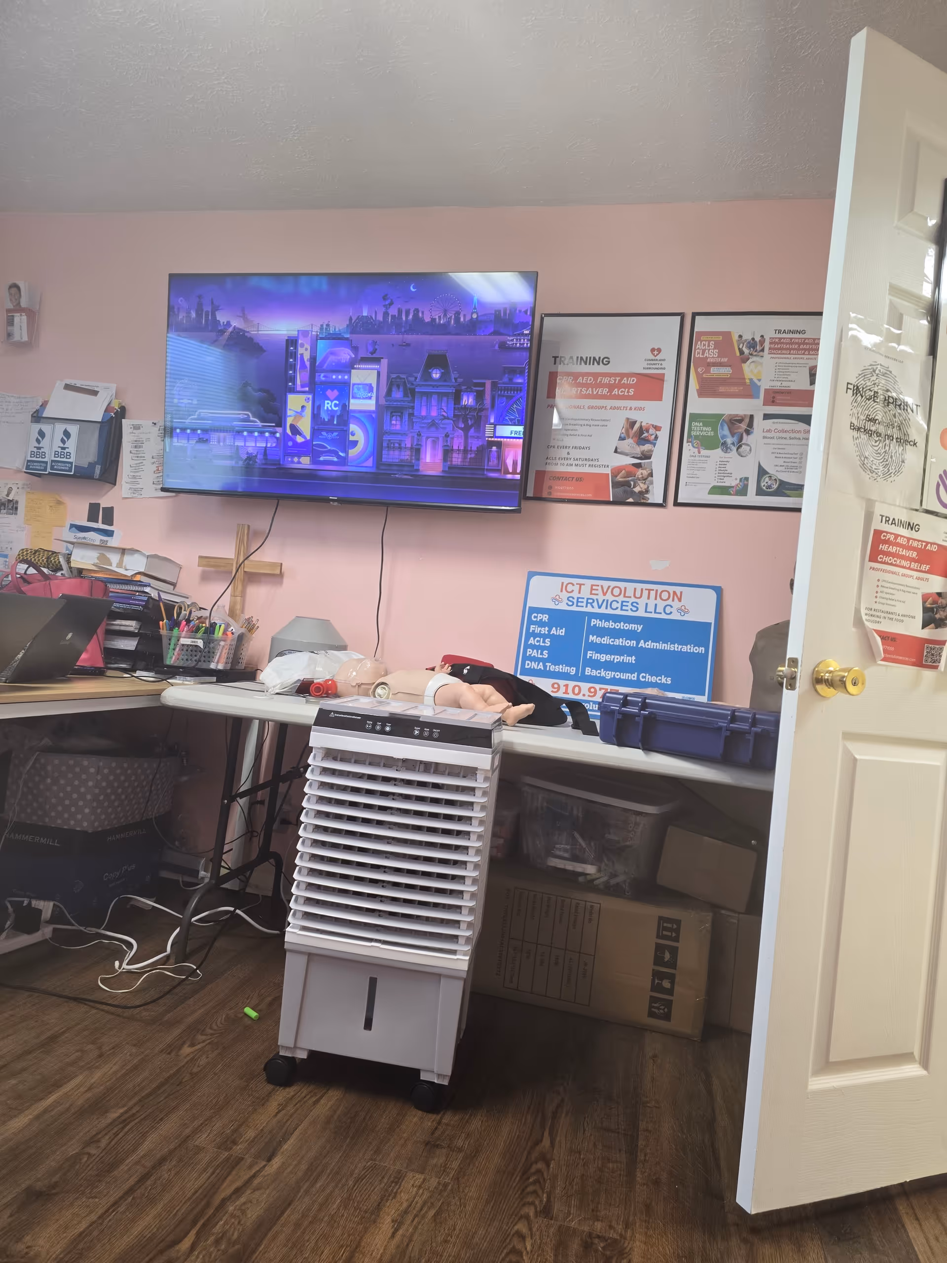 Training room in a facility with a wall-mounted TV, posters, desk, and a CPR dummy.
