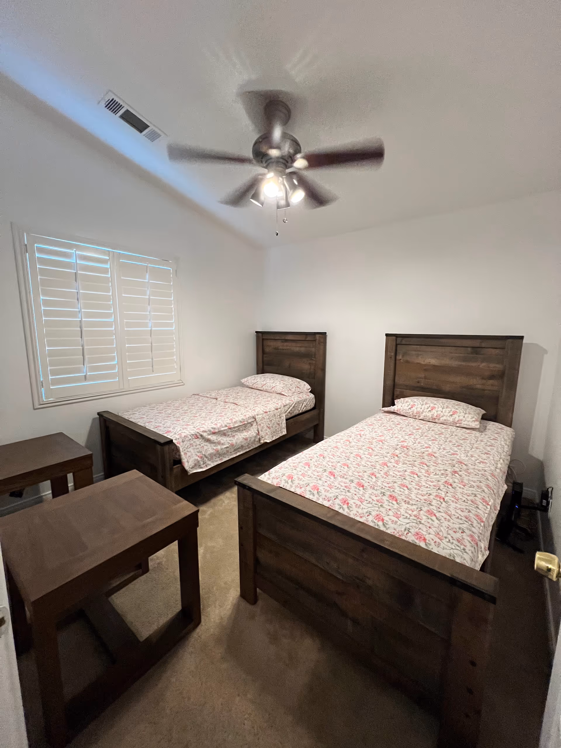 Two single beds with matching wooden frames, nightstands, window blinds, and a ceiling fan.