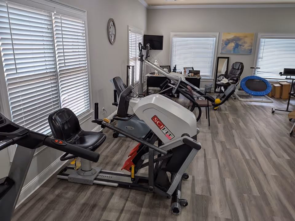 Indoor rehab gym with exercise machines and windows in a senior living facility.