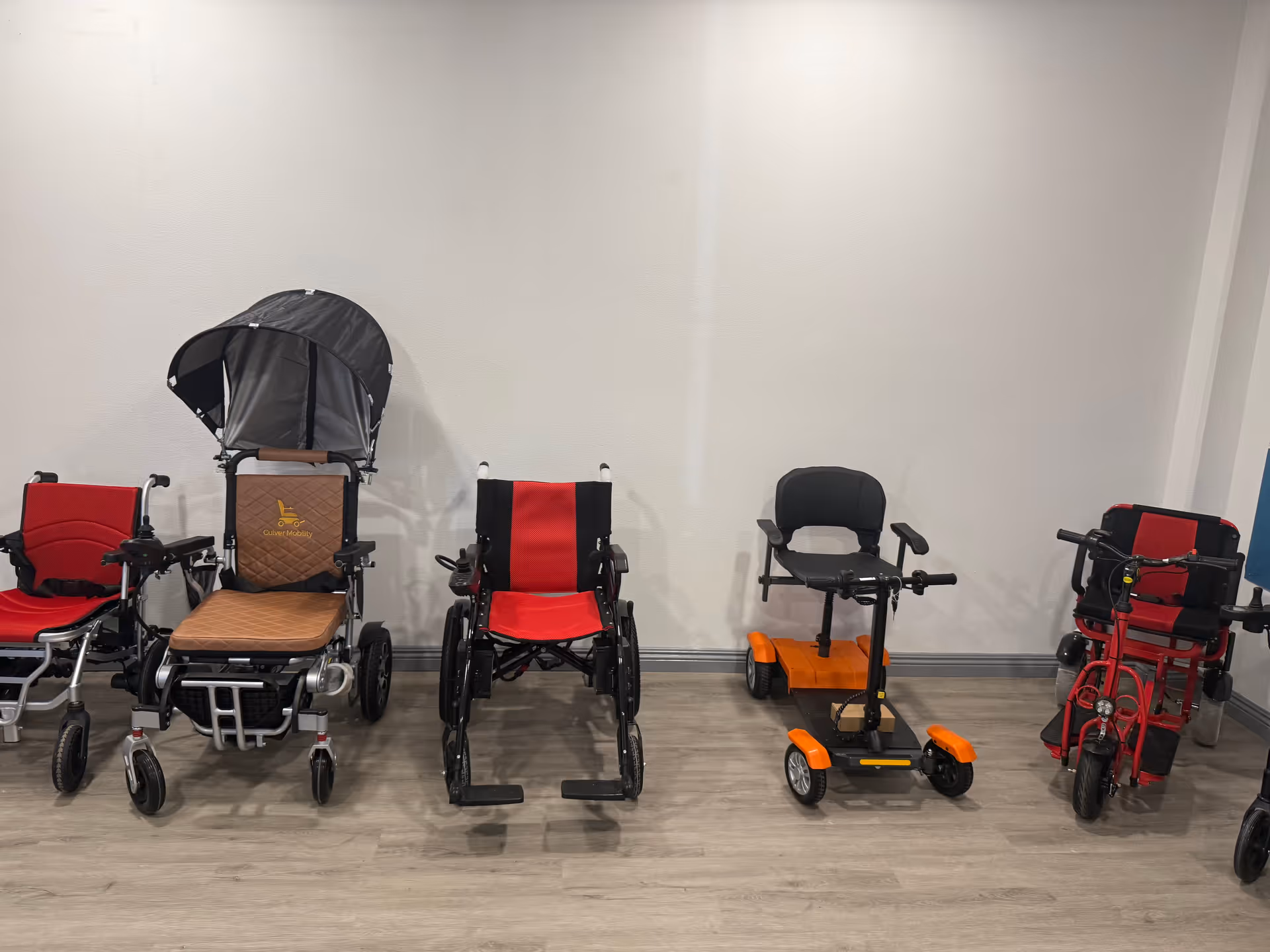 Row of mobility aids and wheelchairs lined up against a plain interior wall.