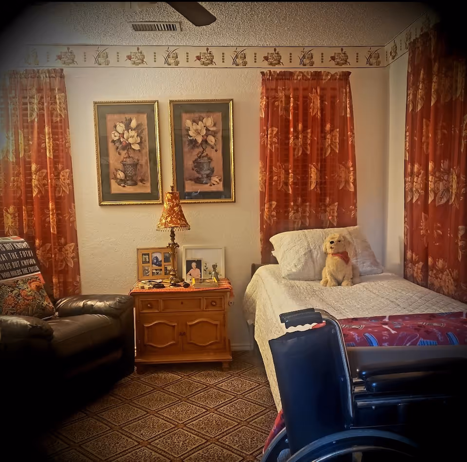 Cozy bedroom in a senior care home with bed, wheelchair, couch, and floral curtains.