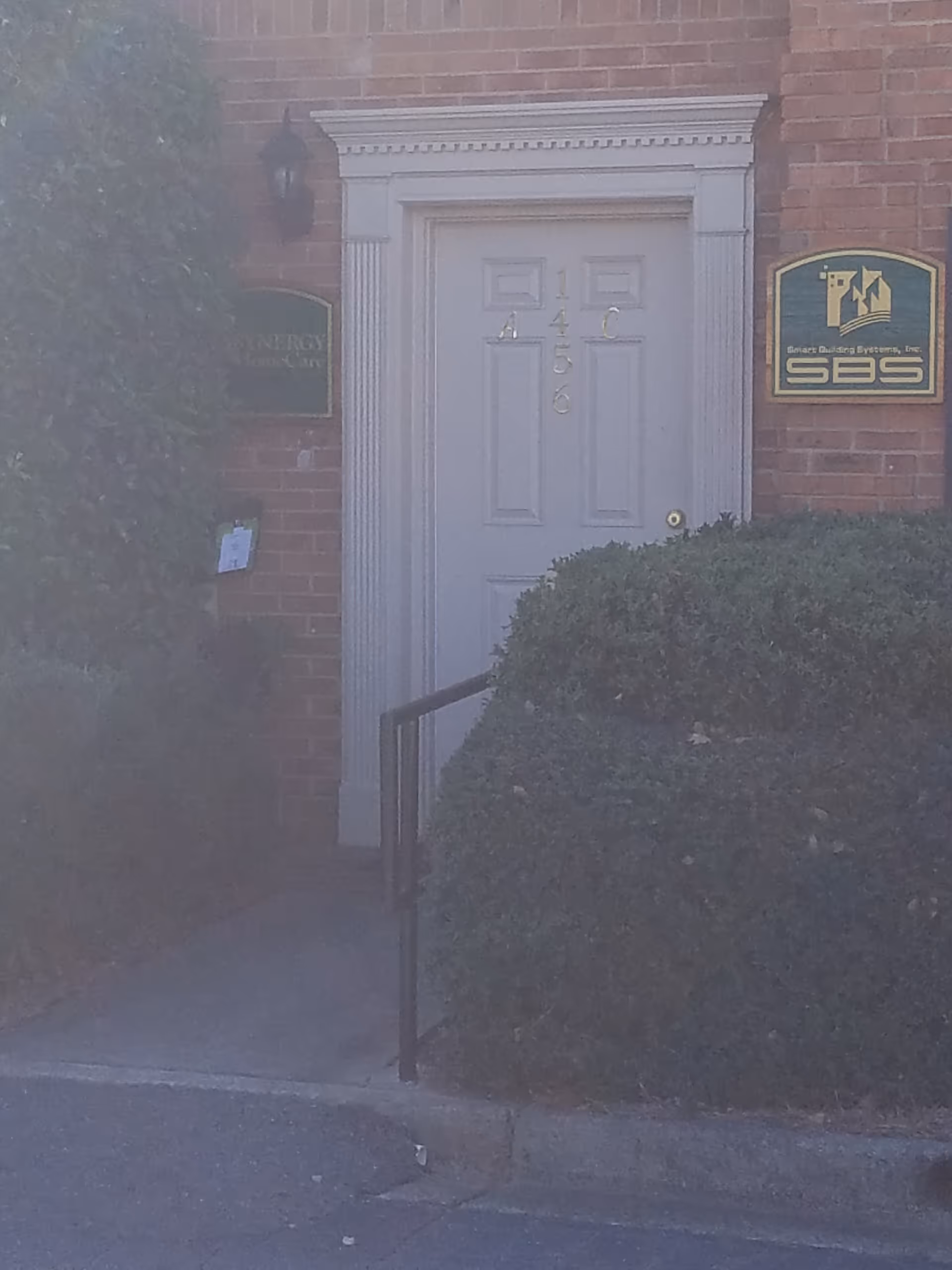 Brick building entrance with a white door, signs, and trimmed hedges.