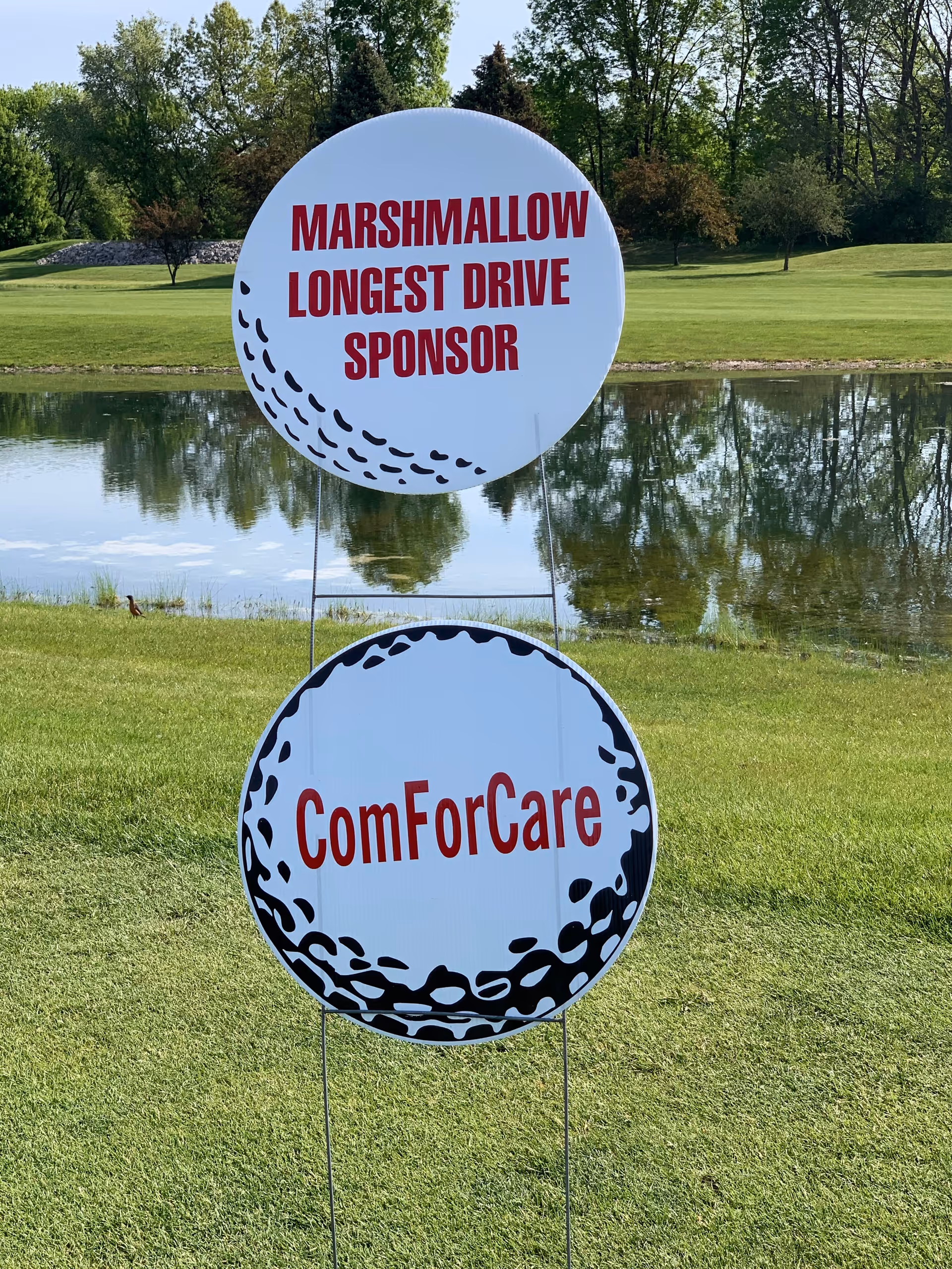 Two round sponsor signs on a grassy golf course beside a reflective pond.