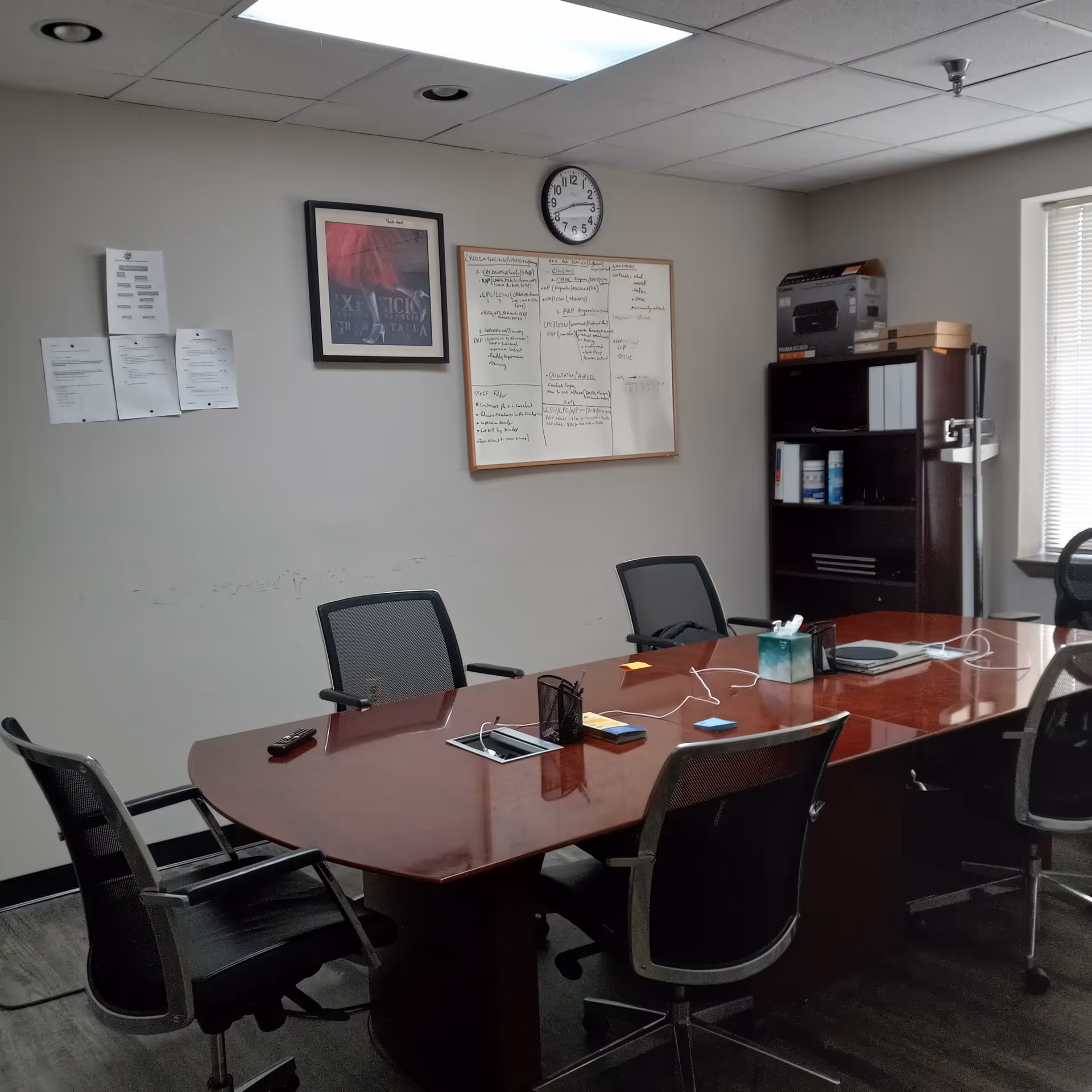 A conference room with a large wooden table, chairs, whiteboard, clock, and office equipment.