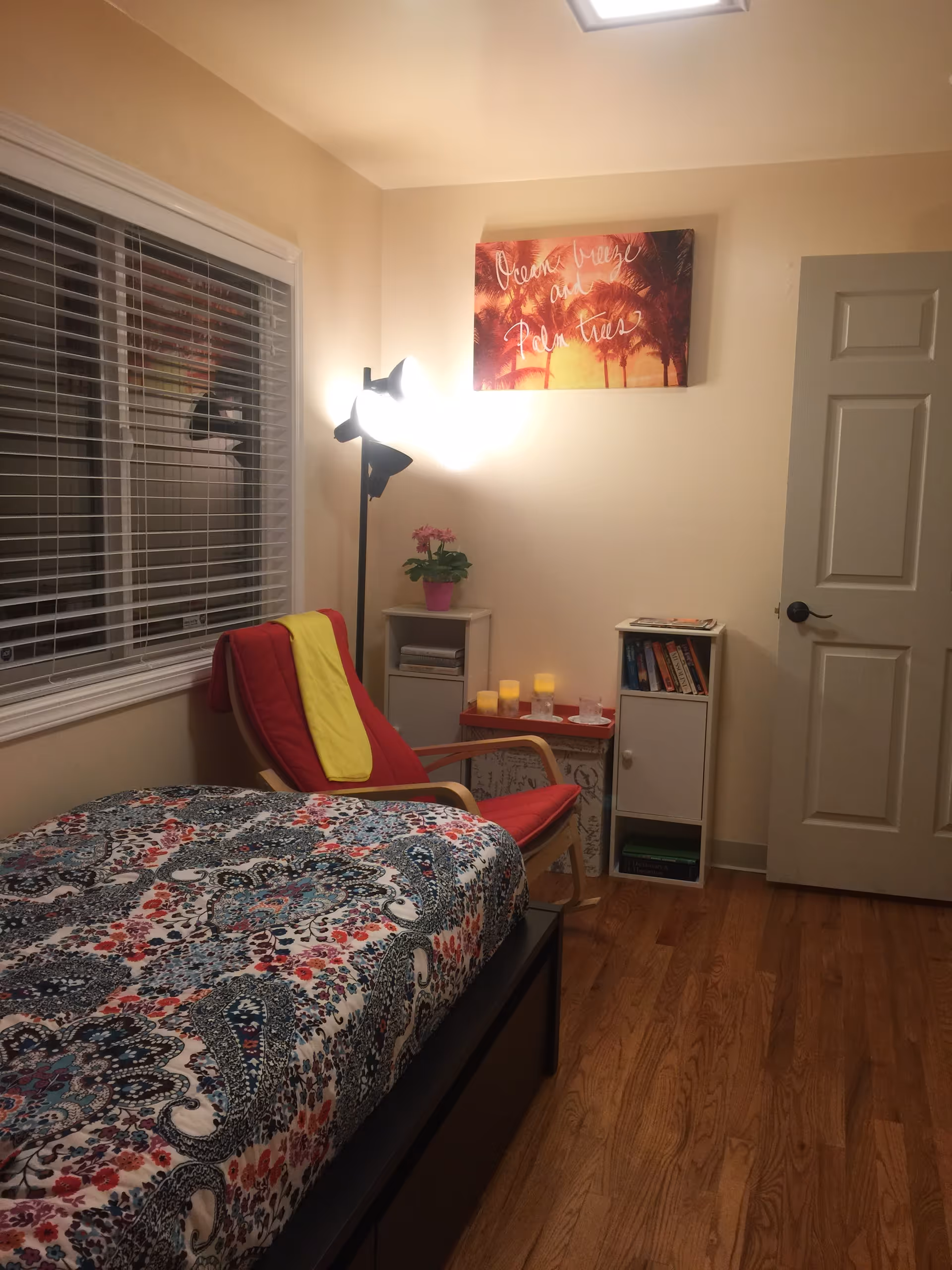 Cozy bedroom with a patterned bed, red chair with yellow throw, plant, candles, and shelves.