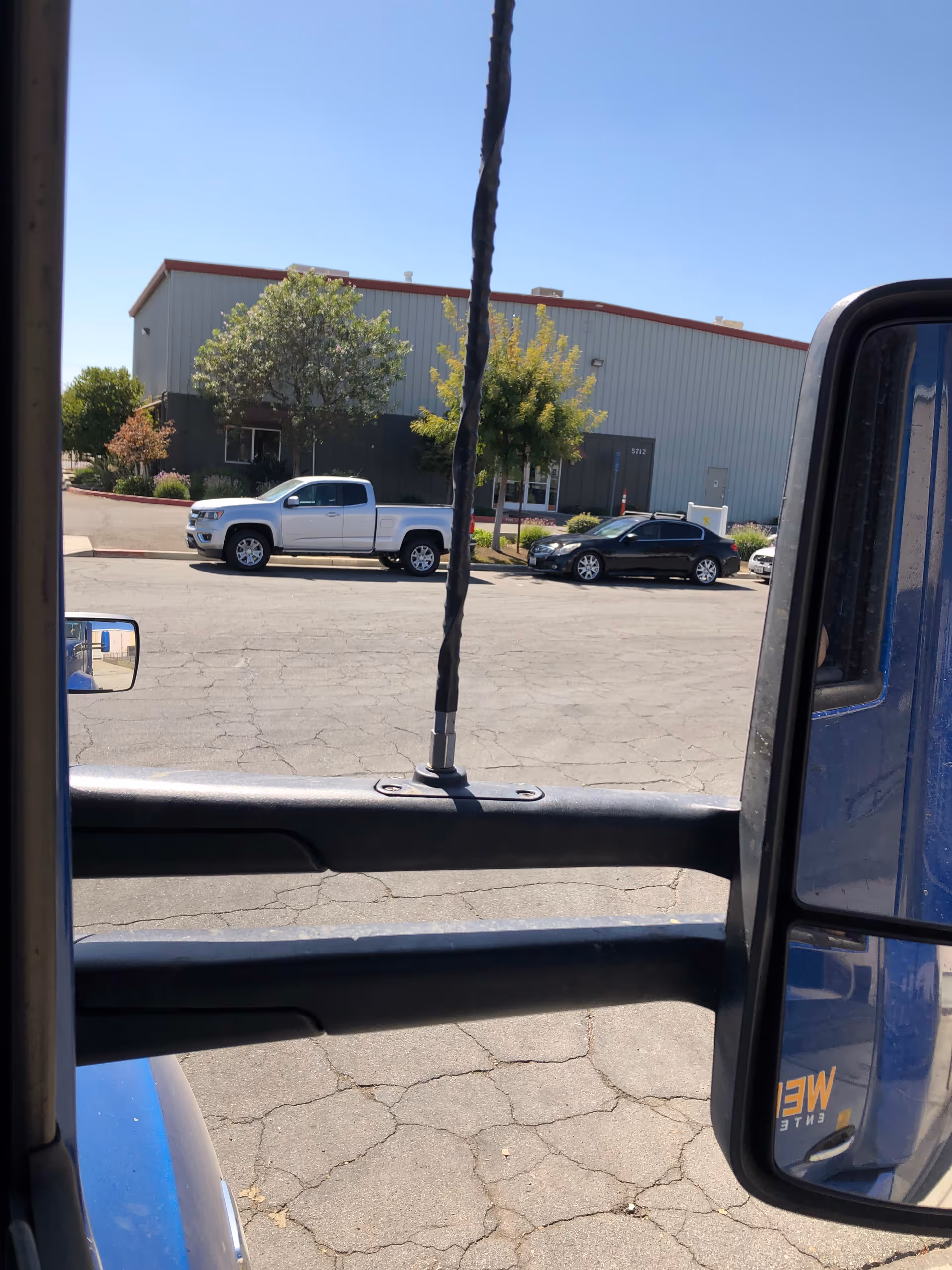 View from inside a vehicle of a parking lot and a gray building with trees.