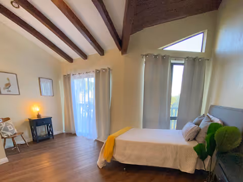 Bright bedroom with vaulted ceiling beams, a bed, curtains, and a potted plant.